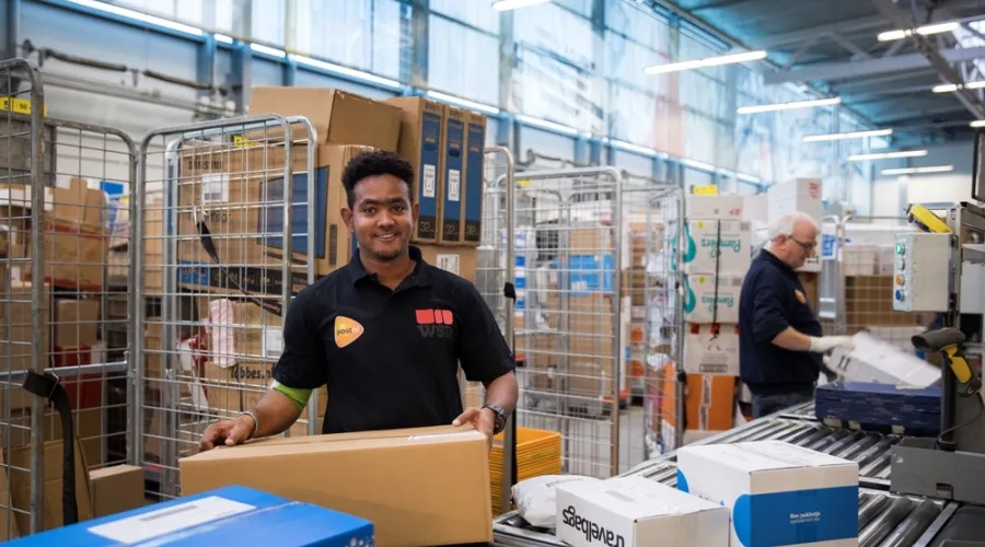 Jonge man lacht in de camera terwijl hij bij PostNL een pakket in zijn hand heeft