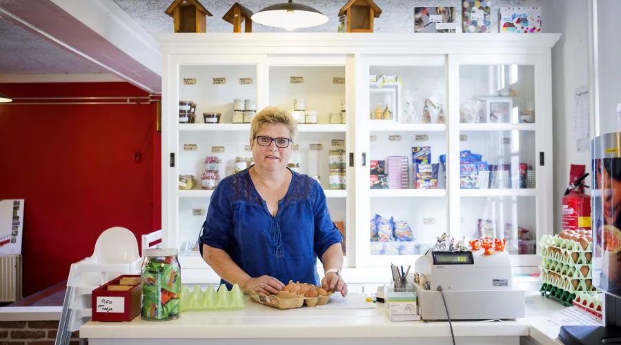 vrouw staat achter de kassa met eieren op de toonbank. Achter haar is een kast met diverse producten te zien