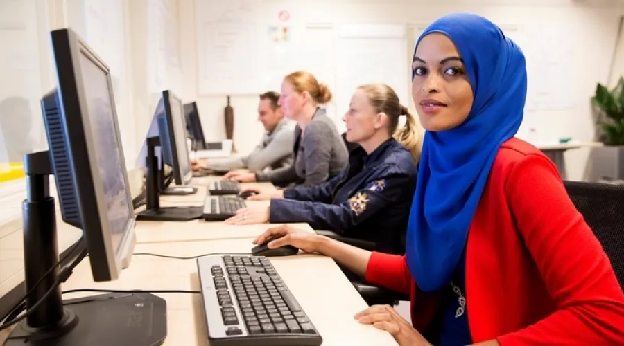 groep mensen in leslokaal zitten achter computers, waarbij een vrouw met hoofddoek vooraan de camera in kijkt