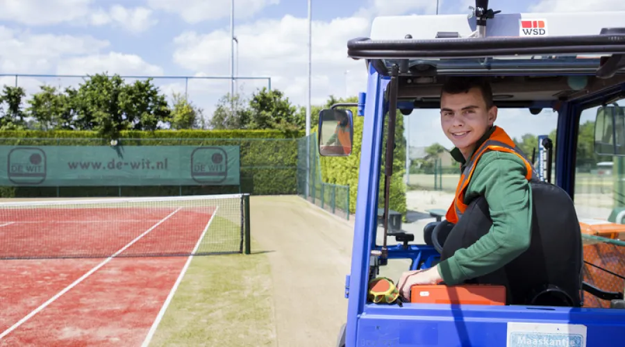 Jongere op een tennisbaan in een heftruck die over zijn schouder naar de camera kijkt