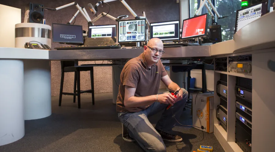 Man zit gehurkt en repareert iets aan een radiostation terwijl hij de camera in kijkt