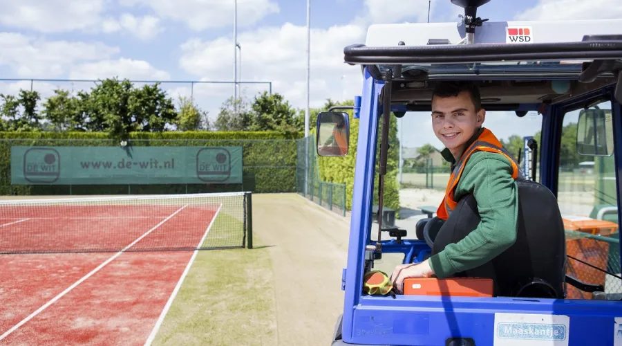 Jongen zit op de heftruck op een tennisbaan en kijkt over zijn schouder lachend de camera in.