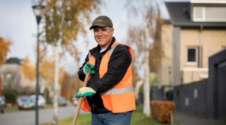 Man die werkt in de groenvoorziening en kijkt lachend in de camera terwijl hij een schoffel vastheeft.