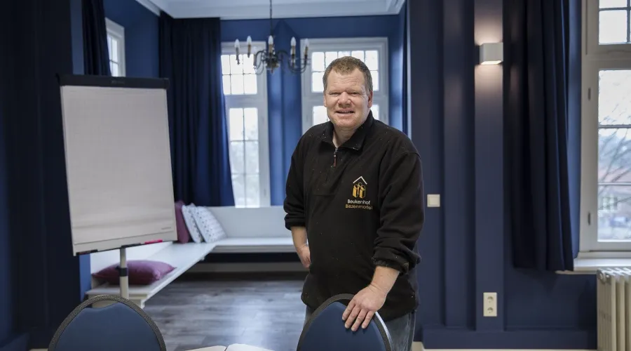 Man staat in een vergaderruimte, leunend met een arm op een stoel en kijkt lachend de camera in.