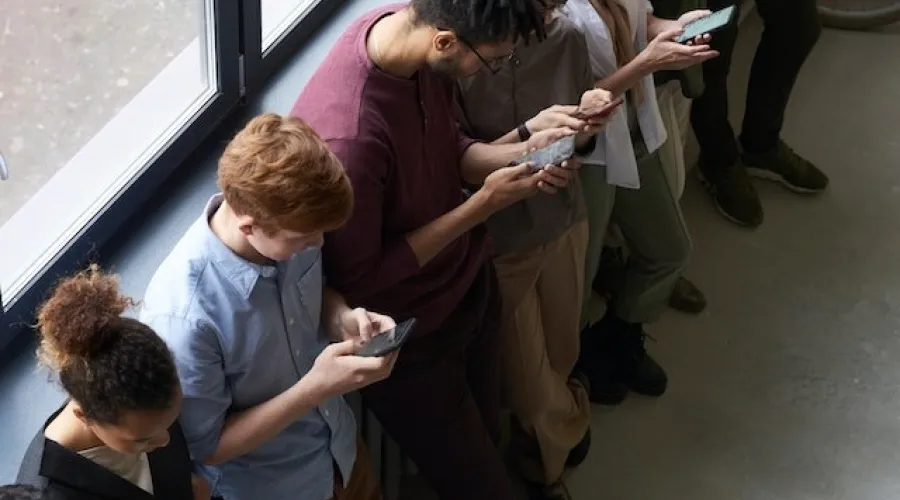 groepje mensen die bij een groot raam op een rij staan en allemaal op hun telefoon in hun handen kijken